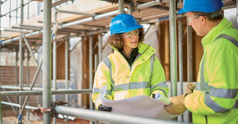 A man and a woman are talking on a construction site. They are wearing yellow reflective coats and blue helmets.