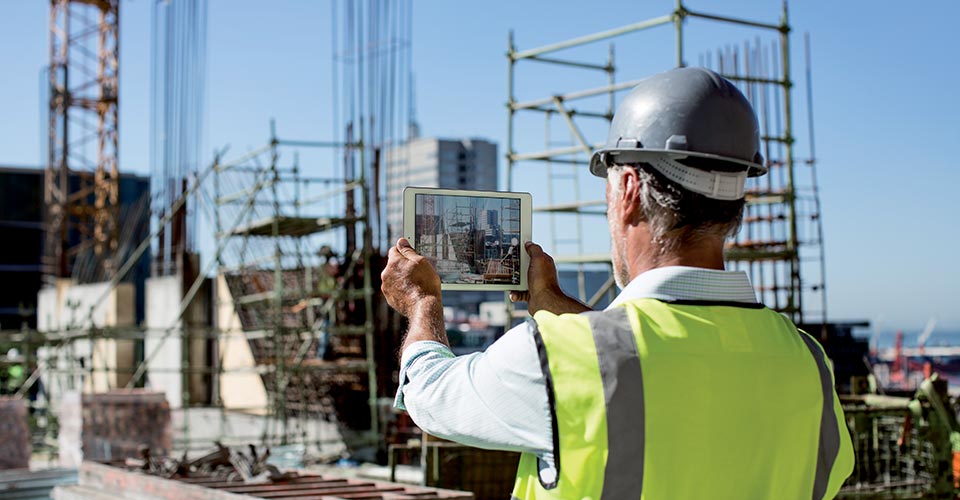 A man is standing in front of a construction site. He holds a tablet in his hands.