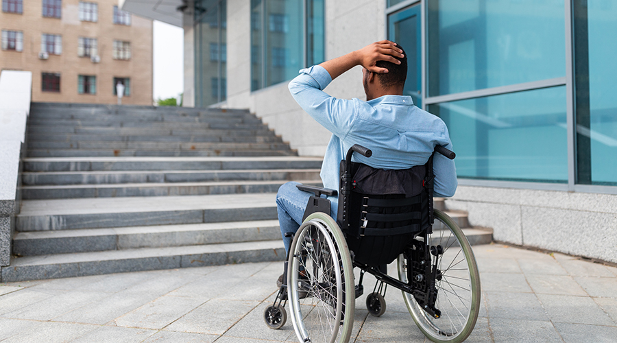 homme en fauteuil roulant qui se tient la tete devant un escalier sans rampe d'accès handicapé